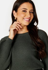 Maglione a coste verde scuro con scollatura rotonda. Orecchino a cerchio in oro con texture che aggiunge un dettaglio. La modella sorride mentre si tocca delicatamente il viso.