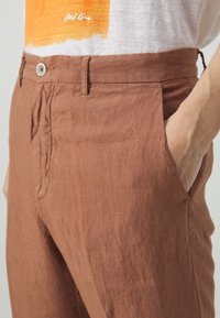 Braune Leinenhose mit entspannter Passform, seitlichen Taschen und glatter Textur, die mit einem Knopfverschluss am Bund geschlossen wird.