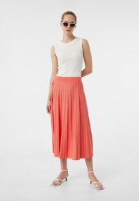 Biała, prążkowana, bez rękawów bluzka zestawiona z plisowaną koralową spódnicą midi z cienkimi białymi paskami. Uzupełniona o białe sandały na ramiączkach.