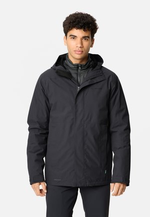 Jonge man met krullend haar die een zwarte winterjas met capuchon en een zwarte broek draagt, staand tegen een effen lichte achtergrond.