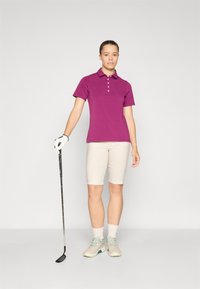 Lila Poloshirt mit kurzen Ärmeln, beigefarbene knielange Shorts, weiße Socken und helle Schuhe. Hält einen Golfschläger. Schlichtes, sportliches Design.