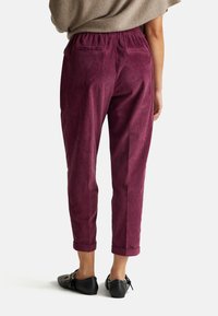 Woman wearing high-waist, rolled-hem, plum-colored corduroy pants and black leather loafers, standing against a plain background.