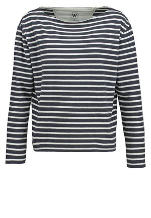 Beige trenchcoat yli navy ja valkoisen raidallisen paidan, yhdistettynä beigeihin leveisiin housuihin ja hopeisiin nauhakengiin. Yksinkertainen, moderni muotoilu.