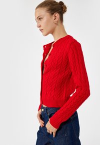 Roter Strickcardigan mit Kabelmuster, rippenbündchen und -säumen, runder Ausschnitt und Knopfverschluss. Trägt man über einer dunklen Jeans.