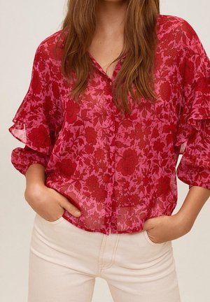 Femme portant un chemisier rouge transparent à fleurs avec des manches volantées et un pantalon beige clair, mains dans les poches.