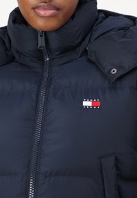 Marineblå pufferjakke med høy krage, glidelås og en liten rektangulær Tommy Jeans-logo med røde, hvite og blå farger.