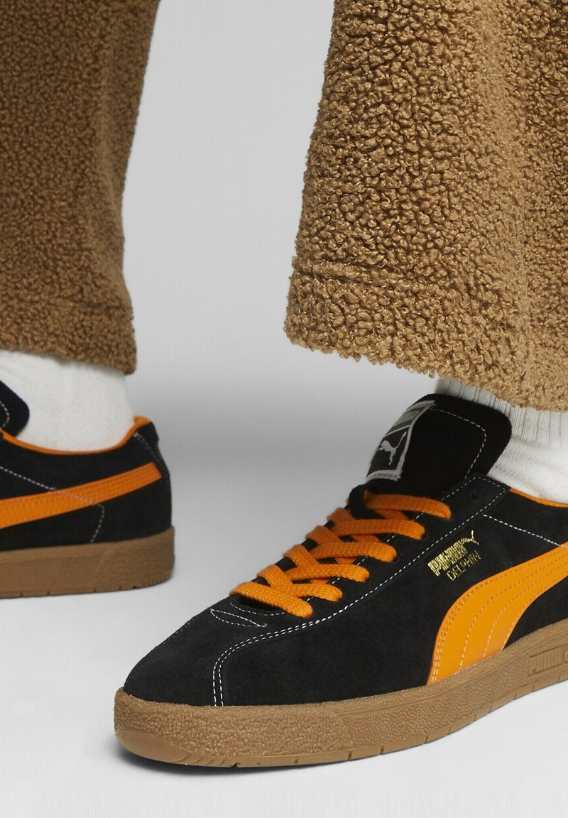 Baskets Puma noires et orange avec semelles marron portées avec des chaussettes blanches et un pantalon marron texturé sur fond gris.