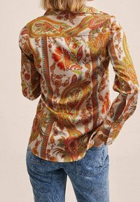 Femme portant une chemise à manches longues avec des motifs floraux paisley orange, rouges et verts, et un jean à motifs paisley bleus, les mains dans les poches arrière.