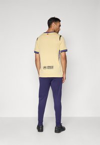 Beige sportshirt met blauwe accenten en UNHCR-logo op de achterkant, gecombineerd met marineblauwe broek en zwarte sneakers. Gladde stofstructuur.