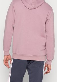 Růžová mikina s kapucí, žebrovanými manžetami a lemem. Tkanina je měkká a hladká. Kombinovaná s tmavě šedými kalhotami s bílými bočními pruhy.