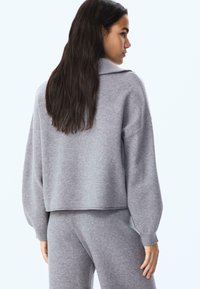 Grau gestrickter Pullover mit hohem Kragen und überschnittenen Schultern, ausgestattet mit gerippten Bündchen. Die Textur wirkt weich und leicht flauschig.