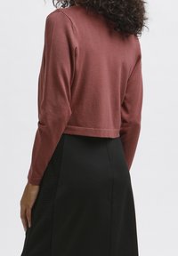 En person med lockigt hår bär en kortärmad, rostfärgad tröja och en svart kjol, vänd bort från kameran mot en vit bakgrund.