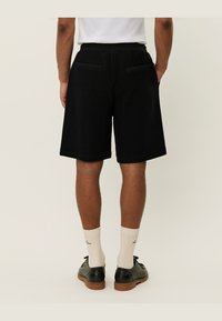 Shorts en mesh noirs avec une finition texturée, dotés de deux poches arrière et d'une taille élastique, associés à des chaussettes couleur crème et des chaussures noires.