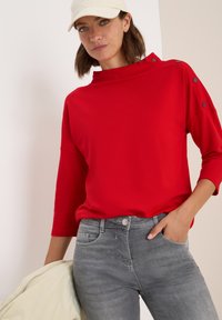 Haut rouge à manches longues avec un col montant et des accents de boutons, associé à un jean gris. Le tissu semble lisse et ajusté.