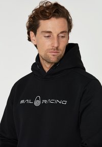 Sudadera negra con capucha ajustable y logotipo bordado "SAIL RACING" en blanco. Textura suave con puños y dobladillo acanalados.