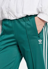 Tīra zila bikses, kas izgatavotas no gludas auduma, ar baltām svītrām, ar rāvējslēdzēja kabatu un Adidas logotipu zemā kājas daļā.