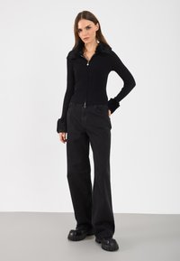 Czarny sweter z prążkowaną strukturą, zapinany na zamek, z kołnierzem i mankietami z sztucznego futra, w połączeniu z wysokim czarnym spodniami o szerokich nogawkach i masywnymi czarnymi butami na platformie.