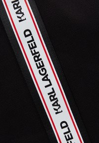 Svart texturerat tyg med en vit rand kantad i rött, där den upprepade texten "KARL LAGERFELD" visas i fet svarta bokstäver.