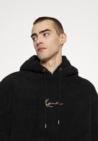 Svart fleece hettegenser med teksturert overflate, justerbar snor i svart med gullakenter, og gulllogo på framsiden.