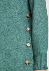 Pulover tricotat de culoare albastru teal, cu manșete cu model  rib pentru o potrivire confortabilă și patru nasturi rotunzi de culoare maro pe lateral. Textură moale cu un model ușor melanj.