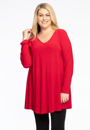 Rote Longsleeve-Tunika mit V-Ausschnitt, fließendem Stoff und lockerer Passform, kombiniert mit schwarzen engen Hosen. Glatte Textur ohne sichtbare Muster.