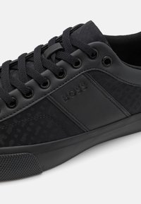 Svart sneaker med en blandning av slät och texturerad material, svarta snören, en mönstrad sidopanel och en gummisula. "BOSS" präglat.