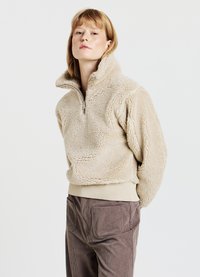 Maglione in pile beige con collo alto e zip a metà, caratterizzato da tessuto strutturato e vita elastica. Abbinato a pantaloni di velluto a coste marroni.