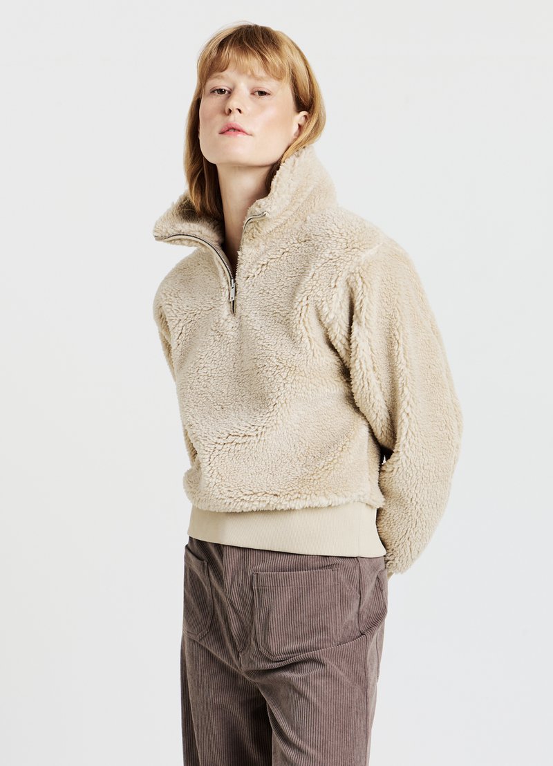 Maglione in pile beige con collo alto e zip a metà, caratterizzato da tessuto strutturato e vita elastica. Abbinato a pantaloni di velluto a coste marroni.
