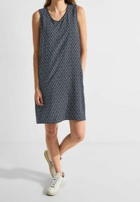 Ärmelloses Kleid in Dunkelblau mit einem Diamantmuster, ausgestattet mit einem V-Ausschnitt und einer lässigen Passform. Kombiniert mit weißen Freizeit-Sneakern.