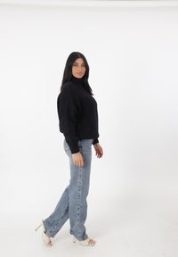 Zwarte gebreide trui met een hoge kraag, gecombineerd met lichtblauwe rechte jeans en beige open tenen hakken. Eenvoudige witte achtergrond.