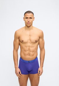 Μυώδης άνδρας στέκεται μπροστά σε λευκό φόντο φορώντας μπλε μπλεκάκια Calvin Klein, κοιτάζοντας προς τα εμπρός με ουδέτερη έκφραση.