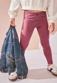 Roza, tesne prilegajoče legice z teksturiranim zaključkom, v kombinaciji z denim jakno in belimi supergami. Ozadje vsebuje nežne pastelne barve.