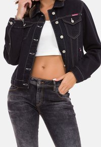 Schwarze Jeansjacke mit auffälliger weißer Naht, silbernen Druckknöpfen und zwei Brusttaschen. Kombiniert mit dunklen Jeans mit einem orangefarbenen Akzent.