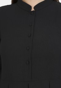 Schwarzes Kleid aus strukturiertem Stoff, mit einem Mandarin-Kragen und einer Vorderleiste mit fünf runden Knöpfen. Schlichte, taillierte Form.