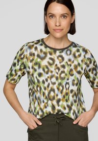 Frau mit kurzen braunen Haaren, die ein kurzärmliges Oberteil mit Leopardenmuster und dunkelgrüne Hosen trägt, mit Händen in den Taschen vor einem grauen Hintergrund.