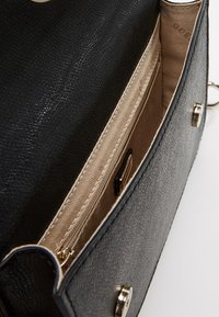 Indvendig i en sort textured lædertaske med en beige foring, der viser en lynlås-lomme og et lille branding-plakette med hardware i guld-tone.