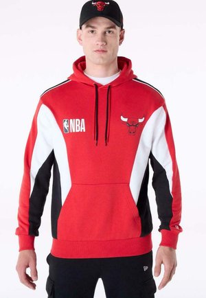 Uomo che indossa una felpa con cappuccio rossa dei Chicago Bulls con il logo NBA e un cappellino nero abbinato con il logo dei Bulls, in piedi davanti a uno sfondo bianco.
