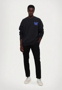 Svart sweatshirt med blå logotyp på bröstet, kombinerad med svarta byxor och vita sneakers. Modellen står rak mot en enfärgad bakgrund.