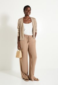 Pantalones beige de pierna ancha con cintura elástica, combinados con una camiseta sin mangas blanca y un cárdigan metálico beige. Incluye un pequeño bolso de mano tejido.