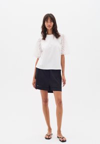T-shirt bianco con maniche a sbuffo in pizzo, abbinato a pantaloni corti neri con orlo decorativo. Il modello indossa ciabatte nere.