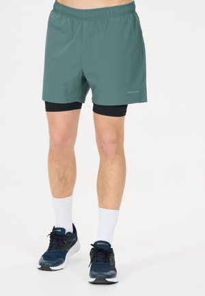 Mandeben iført grønne atletikshorts med sorte kompressionsshorts under, hvide ankelstrømper og mørkeblå løbesko.
