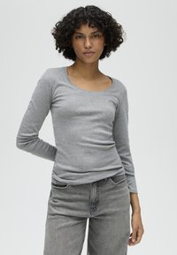 Giovane con capelli ricci che indossa una maglia grigia aderente a maniche lunghe e jeans grigi, in piedi con una mano dietro la schiena.