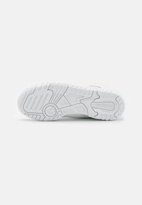 Semelle d'une basket blanche New Balance montrant les motifs de la semelle et le texte "non-marking sole" sur un fond clair uni.
