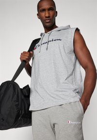 Grijze mouwloze hoodie met een "Champion"-logo, gecombineerd met grijze joggingbroek met een geborduurd logo. Zwarte sporttas met schouderband.