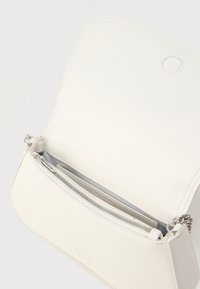 Λευκή δερμάτινη τσάντα με απαλή υφή, τσέπη με φερμουάρ και στρογγυλεμένο επάνω καπάκι. Διαθέτει ασημένια μεταλλικά στοιχεία και διακριτικές ραφές.