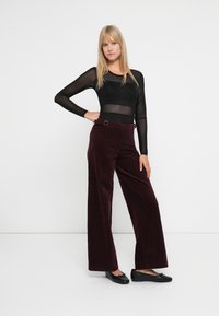 Femeie stând cu mâna pe șold, purtând un top lung și transparent de culoare neagră, pantaloni largi de culoare burgundă închisă și balerini negri, pe un fundal alb.
