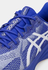 Zapatilla deportiva azul con una parte superior de tela texturizada, que presenta un logo y acentos blancos, y un sistema de cordones reforzado para mayor soporte.