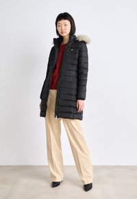 Must ahi-toppmantel kapuutsiga, mille ääres on karvadega ääristus, luku sulgemine ja küljetaskud. Ühildatud punase kampsuni ja beežide laiade pükstega.