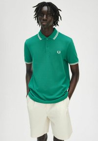 Polo vert avec col et rayures blanches aux manches, arborant un petit logo sur la poitrine gauche. Porté avec un short de couleur claire.