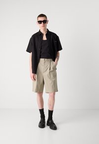 Camisa negra de manga corta sobre una camiseta negra sin mangas, combinada con pantalones cortos beige. Los accesorios incluyen gafas de sol negras, calcetines gruesos y zapatos negros.